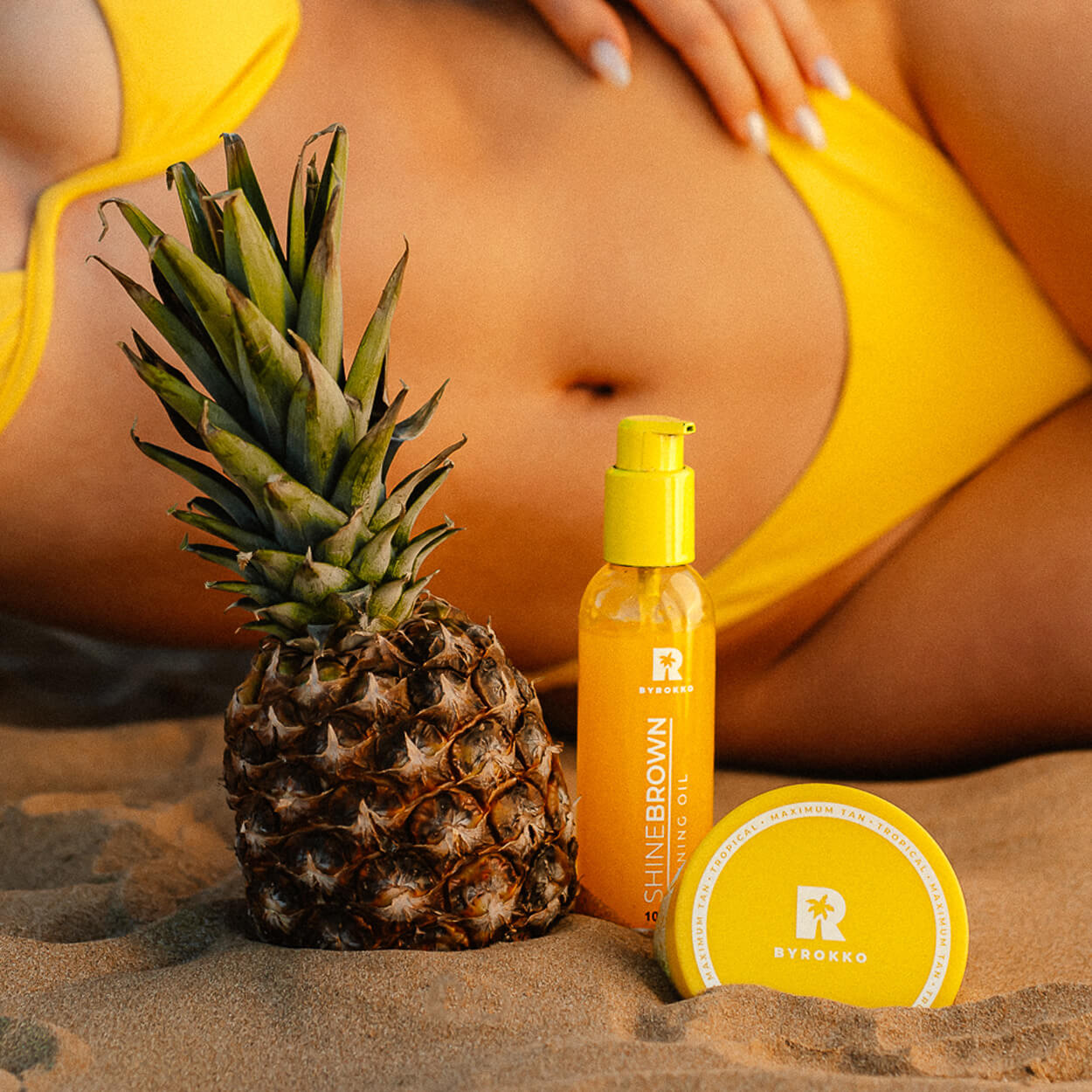 Byrokko Shine Brown Tropical tanning cream and tanning oil on the sand, beside a pineapple and a girl in a yellow bikini.